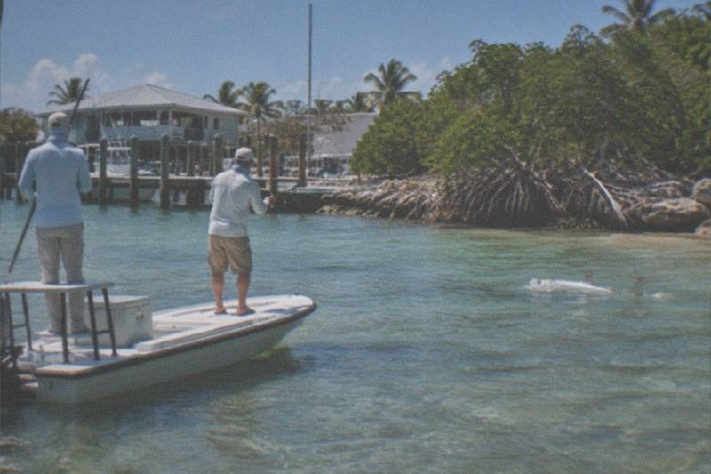 A flats skiff gliding across shallow backcountry water near Key West mangroves, a guide on the poling platform while an angler casts toward a rolling tarpon