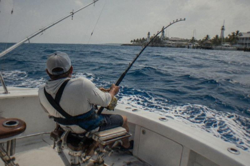 A deep-sea fishing boat under way past the reef line into blue offshore swells, an angler braced at the rail with a heavy rod doubled over under load