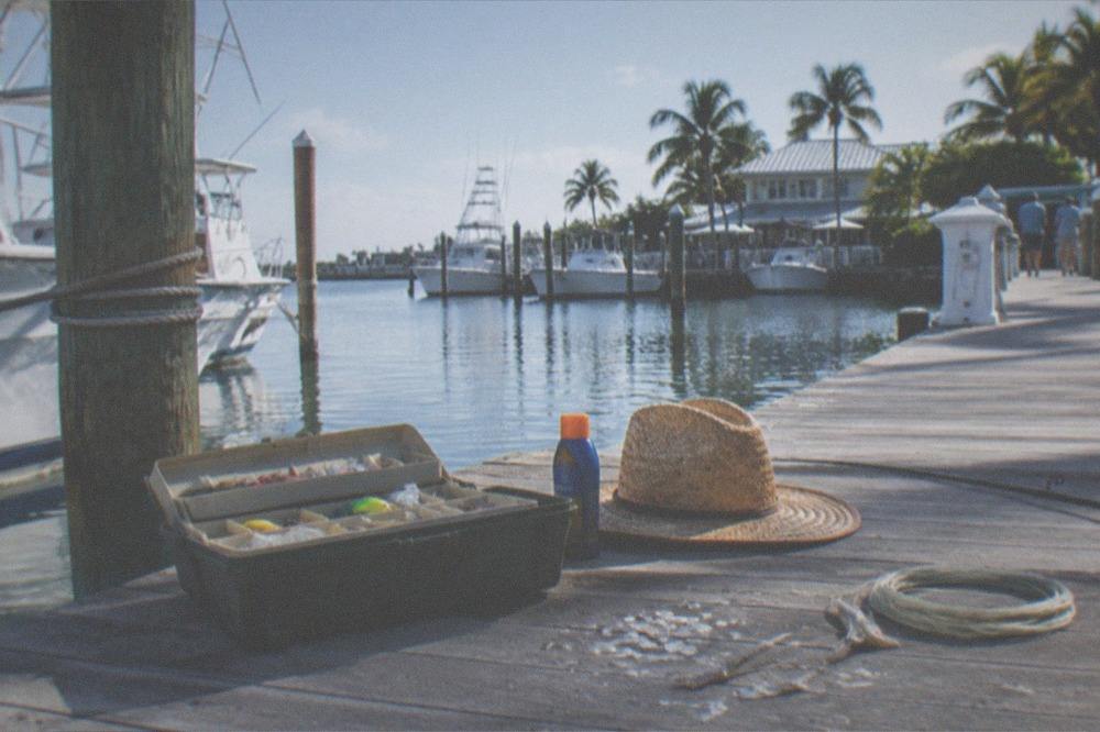 A tackle box, sunscreen bottle, and hat arranged on a wooden dock beside calm water