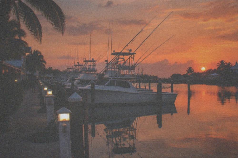 A marina row of charter boats tied up at dusk with outriggers raised against an orange sky